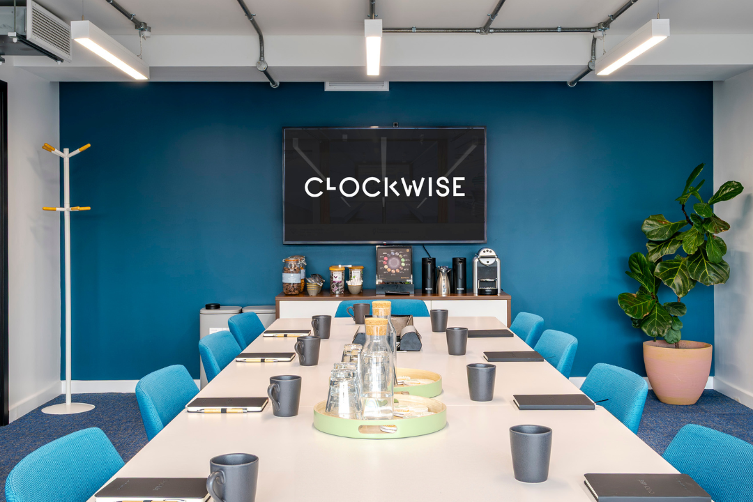 A modern conference room with a long table, blue chairs, a TV screen displaying 