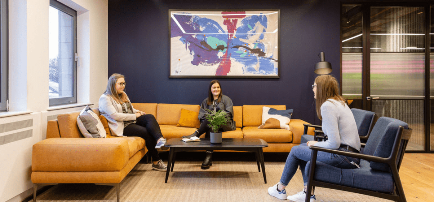 Three people are seated on a tan sofa and blue chairs in a modern lounge with a large abstract artwork on a dark wall. A small coffee table with a potted plant is in the center. Bright windows and a glass door are in the background.