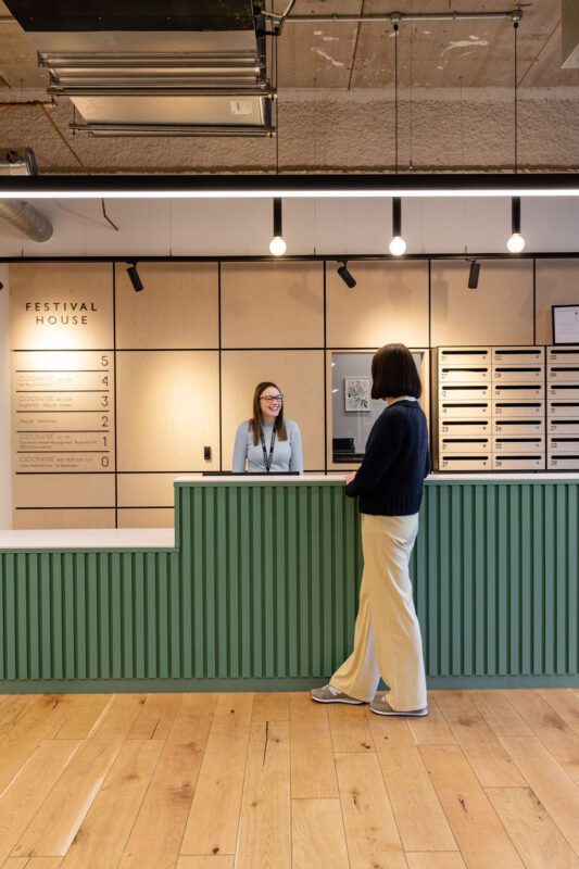 A person stands at a green reception desk speaking to a receptionist. The receptionist smiles from behind the counter. The background features a wall with cubbyholes and a sign reading 