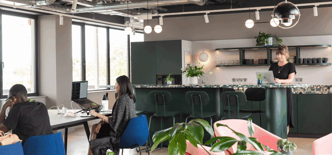 A modern, open-plan office with several people working at desks and a woman standing at a laptop near a kitchen counter. Large windows, plants, and contemporary lighting decorate the space.