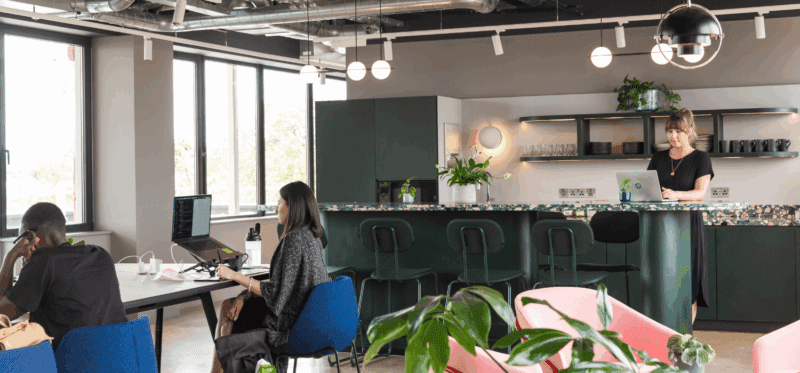 A modern, open-plan office with several people working at desks and a woman standing at a laptop near a kitchen counter. Large windows, plants, and contemporary lighting decorate the space.