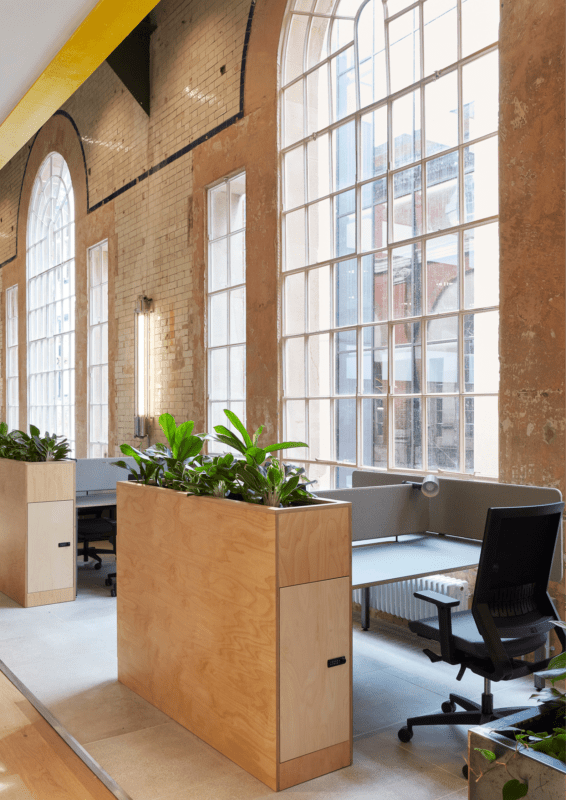 Espace de bureau lumineux avec de hautes fenêtres cintrées et des murs en briques apparentes. Deux bureaux en bois avec des chaises et des plantes vertes sont présentés. La lumière du soleil traverse les fenêtres, mettant en valeur le design moderne et aéré.
