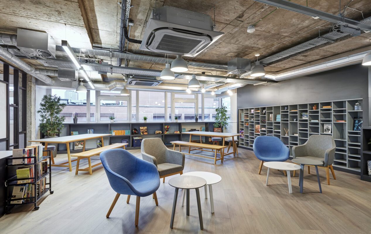 Modern office space with wooden floors, exposed ceilings, and large windows. Features include grey shelving units, blue and grey armchairs, small round tables, long work tables, and potted plants, creating a contemporary and open atmosphere.