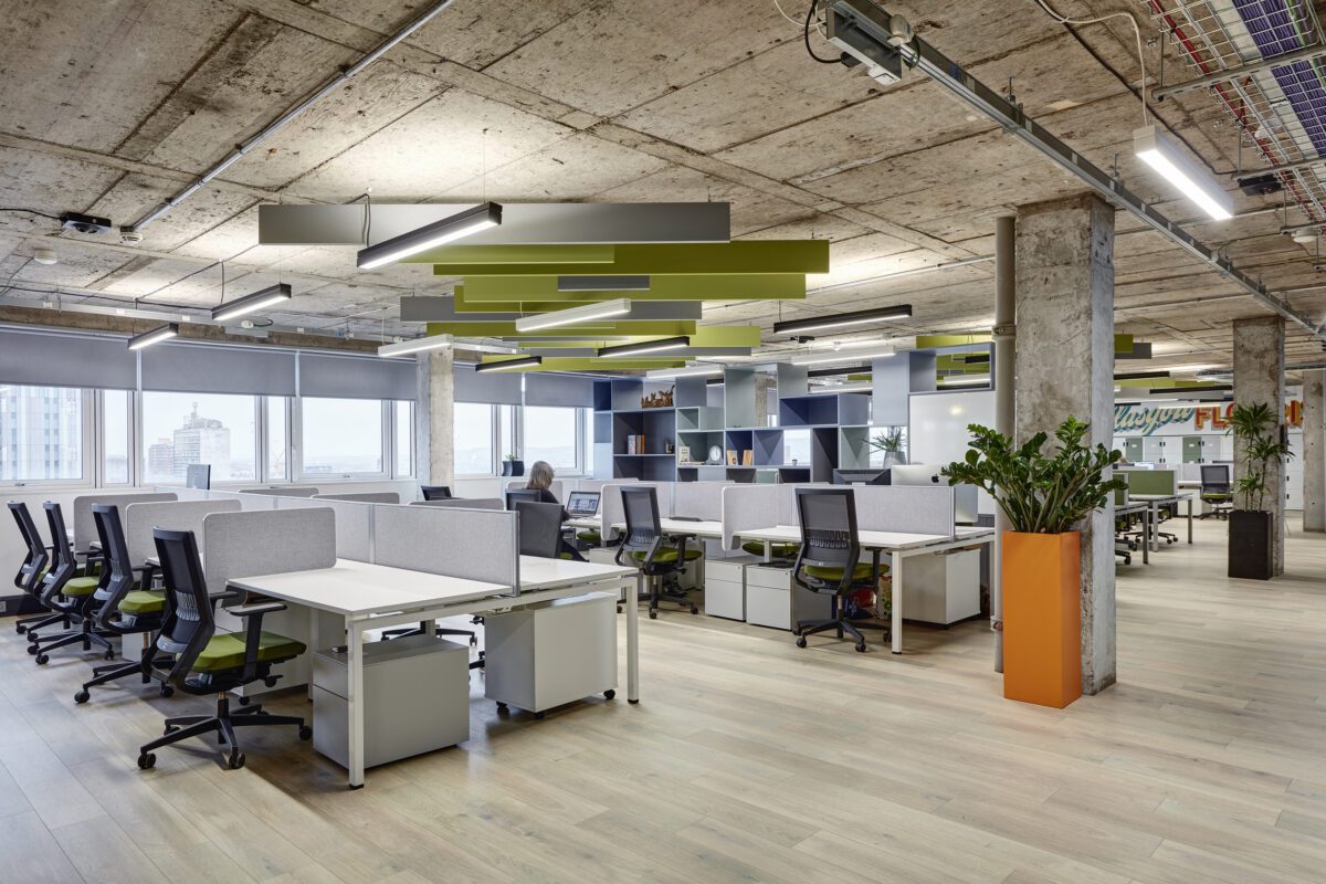 Open-plan office with modern design, featuring white desks and ergonomic chairs. Green and gray ceiling panels add color. Large windows offer city views. Potted plants and an orange planter provide a touch of nature. Ceiling shows exposed concrete.