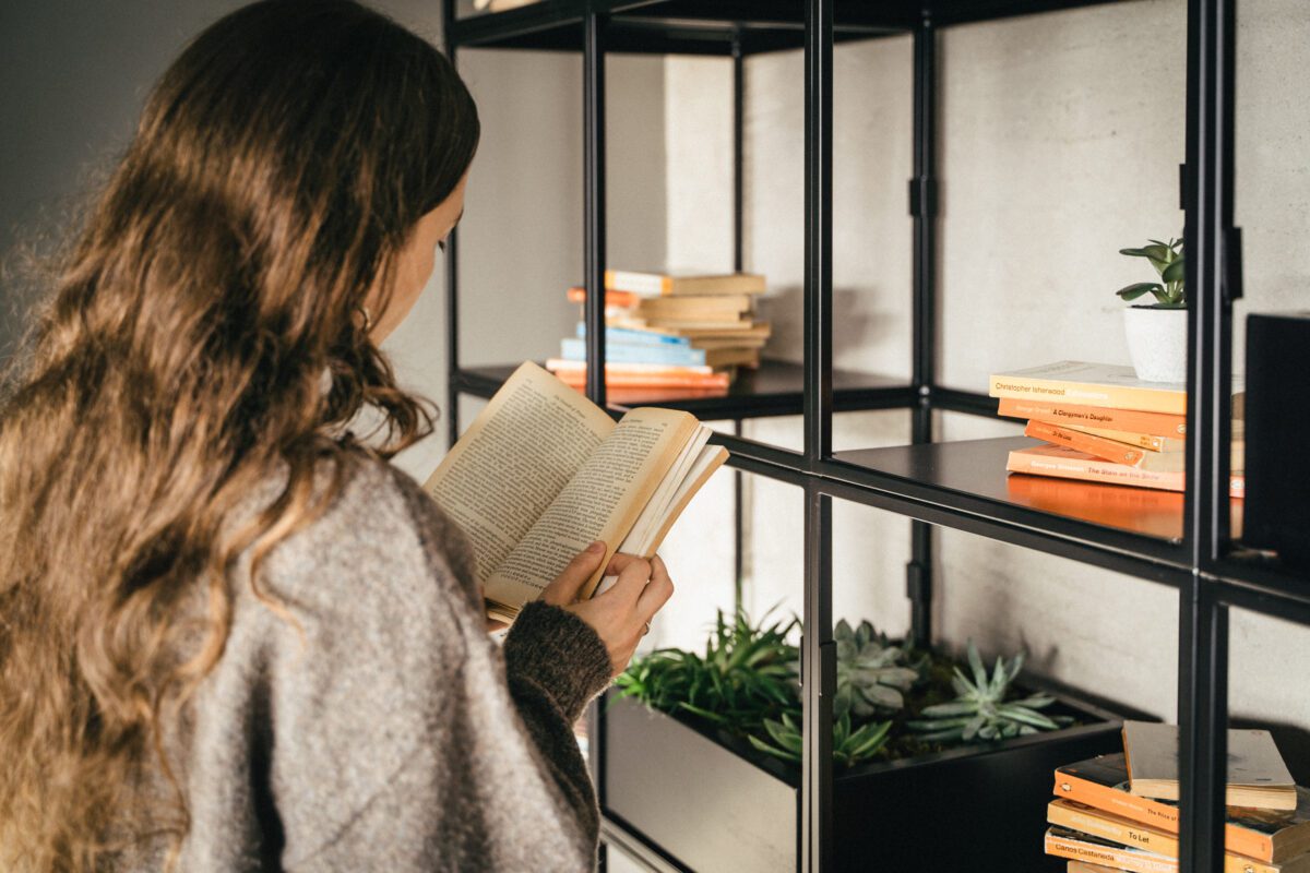 A person with long, wavy hair is reading a book while standing next to a black metal bookshelf. The shelf has several books arranged neatly, along with small potted plants. The wall behind is gray and the lighting is warm.