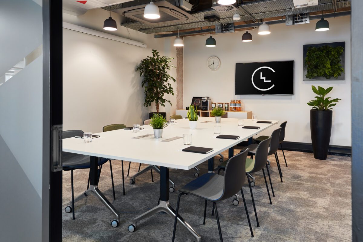 A modern conference room with a long white table, eight chairs, and potted plants. A large TV screen mounted on the wall displays a logo. The ceiling features exposed pipes, and a clock is visible on a shelf with refreshments.