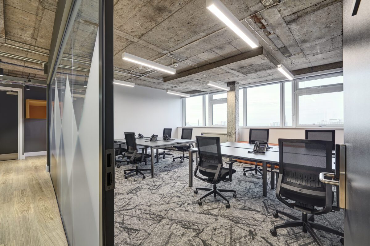 Modern office space with concrete ceiling, glass partition, and six workstations. Each desk has a black office chair and a phone. Large windows allow natural light into the room, and the floor has a patterned carpet.