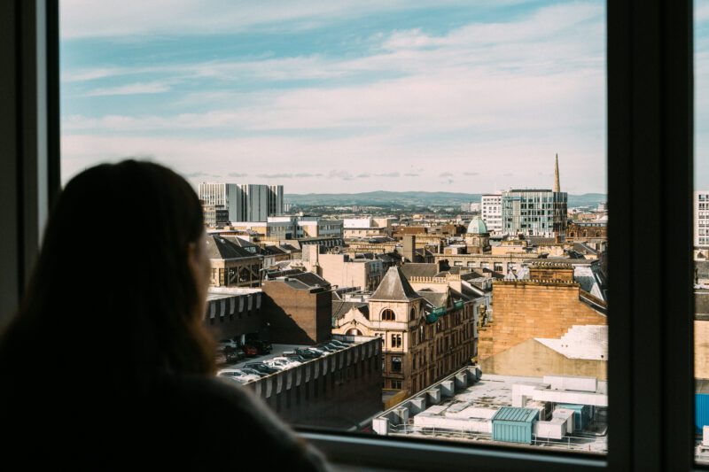 A person looks out of a window at a cityscape under a blue sky. The view includes various buildings with a mix of architectural styles, and hills can be seen in the distance.