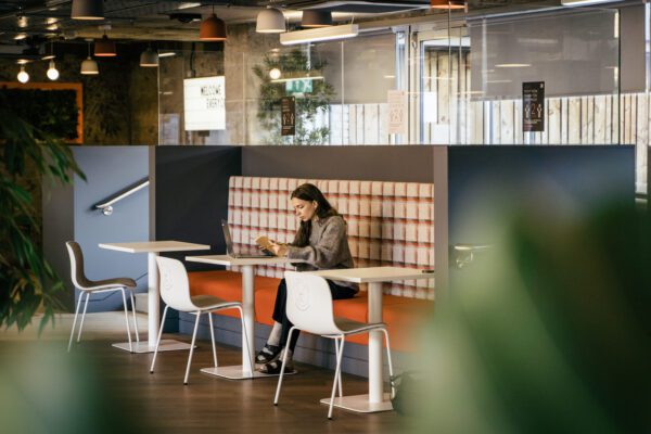 Een vrouw zit alleen aan een tafel in een modern café en werkt op haar laptop. De ruimte beschikt over een gezellige, met ruitjes beklede bank, moderne witte stoelen en warme verlichting. Groene planten omlijsten de scène gedeeltelijk.