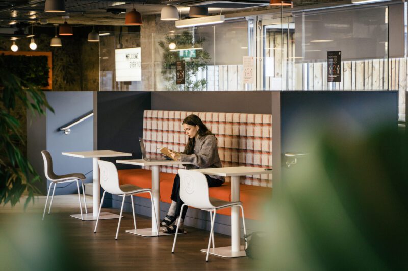 A person sits at a table in a modern cafe with plaid upholstery, working on a laptop. The area is decorated with green plants and stylish lighting. Several empty tables and chairs surround them.