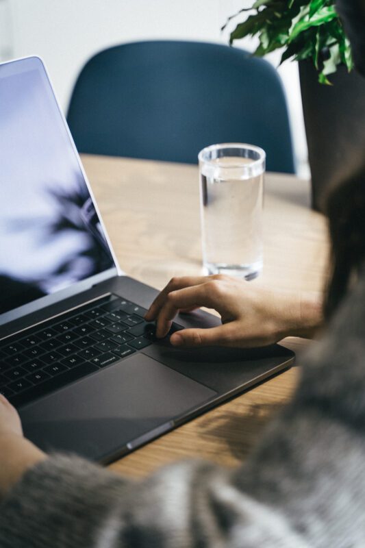 A person using a laptop with a glass of water on a wooden table nearby. Part of a leafy plant is in the background. The setting appears to be a casual work or study environment.