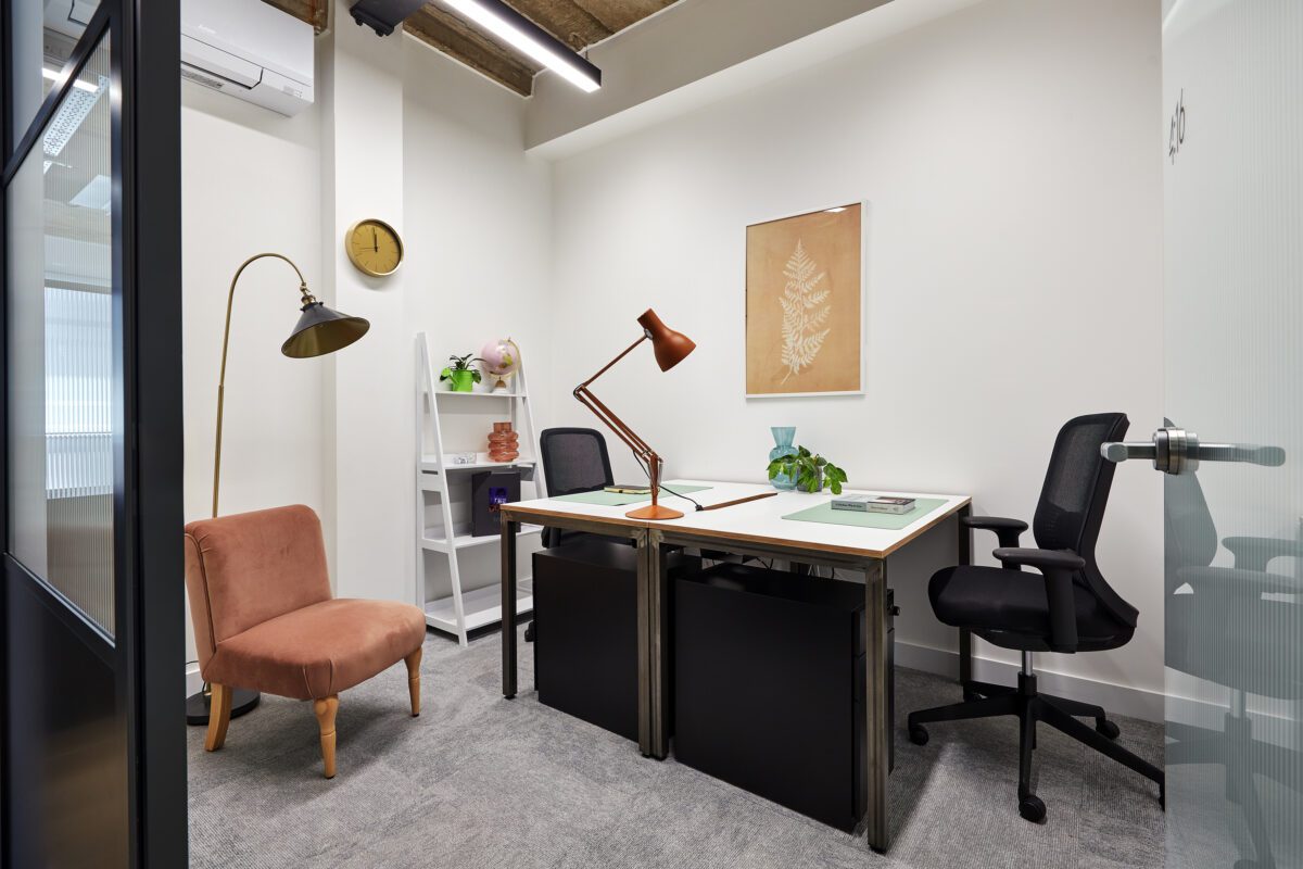 A modern office room with two desks and chairs, a standing lamp, a globe, and a small plant. A framed leaf print hangs on the wall, next to a shelf with books and a clock. A coral-colored armchair is in the corner. The room has gray carpet.
