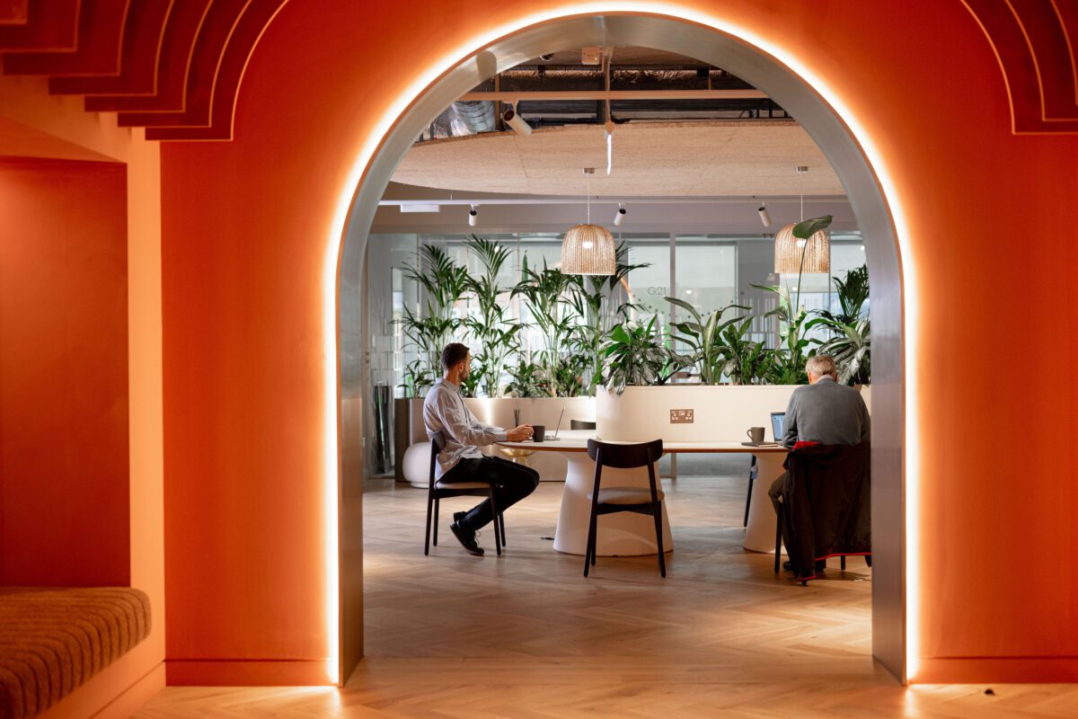 A dimly lit, modern workspace features two people seated at a desk in front of numerous plants. They are framed by a large, glowing archway in a warm-toned room, with wooden flooring and stylish overhead lamps.