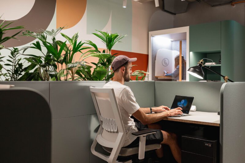 Man in a cap works on a laptop at a modern workstation with green dividers and potted plants. Another person appears in a phone booth in the background. The walls feature abstract designs in muted colors.