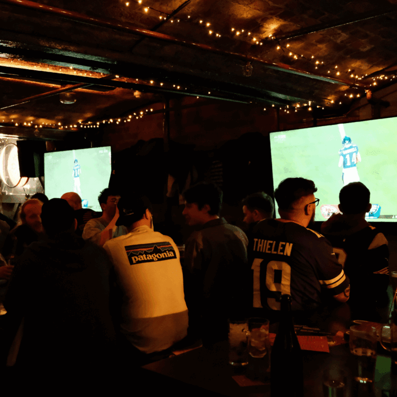 A dimly lit bar with people watching a football game on large TV screens. String lights hang from the ceiling, and several drinks are on the tables. One person is wearing a