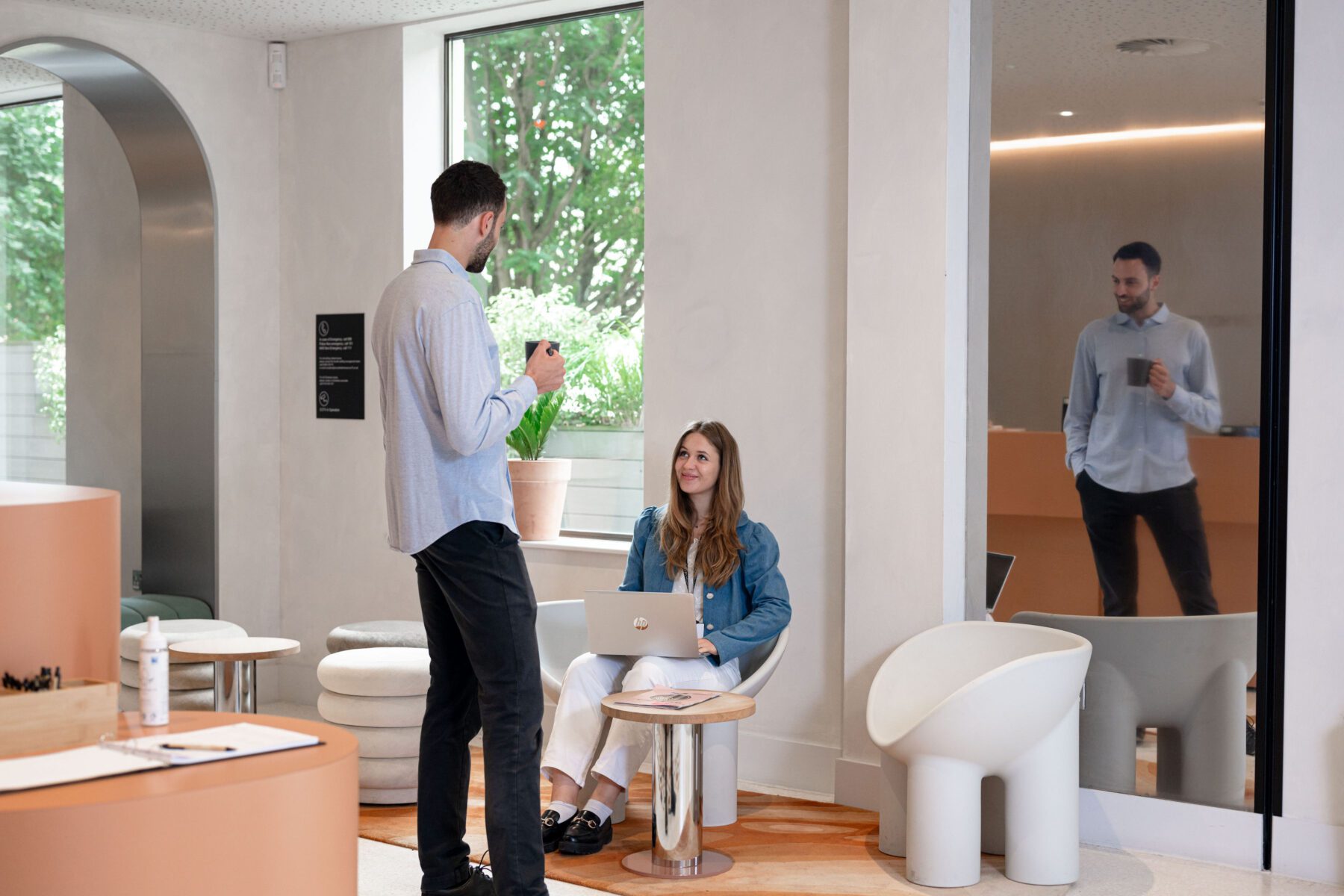 A woman sits on a chair with a laptop on her lap, looking at a man standing in front of her. The man gestures while holding a cup. They are in a modern room with plants, natural light, and a reflection in a large mirror.