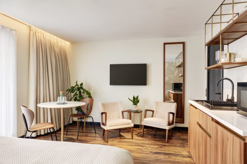 Modernes Hotelzimmer mit Parkettboden, einem Bett, einem runden Tisch mit Stühlen, zwei beigen Sesseln und einem an der Wand montierten Fernseher. Es gibt eine Küchenzeile mit Spüle und Mikrowelle sowie ein großes Fenster mit transparenten Vorhängen, das für Tageslicht sorgt.