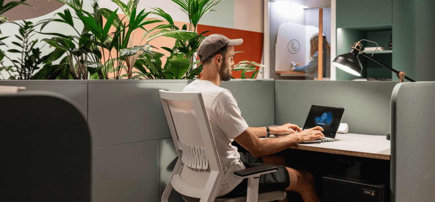 Une personne portant une chemise blanche et une casquette travaille sur un ordinateur portable à un bureau dans un espace de travail. La zone est entourée de cloisons végétales. Une autre personne est vue entrant dans une pièce avec un symbole circulaire sur la porte. L'éclairage est clair et naturel.