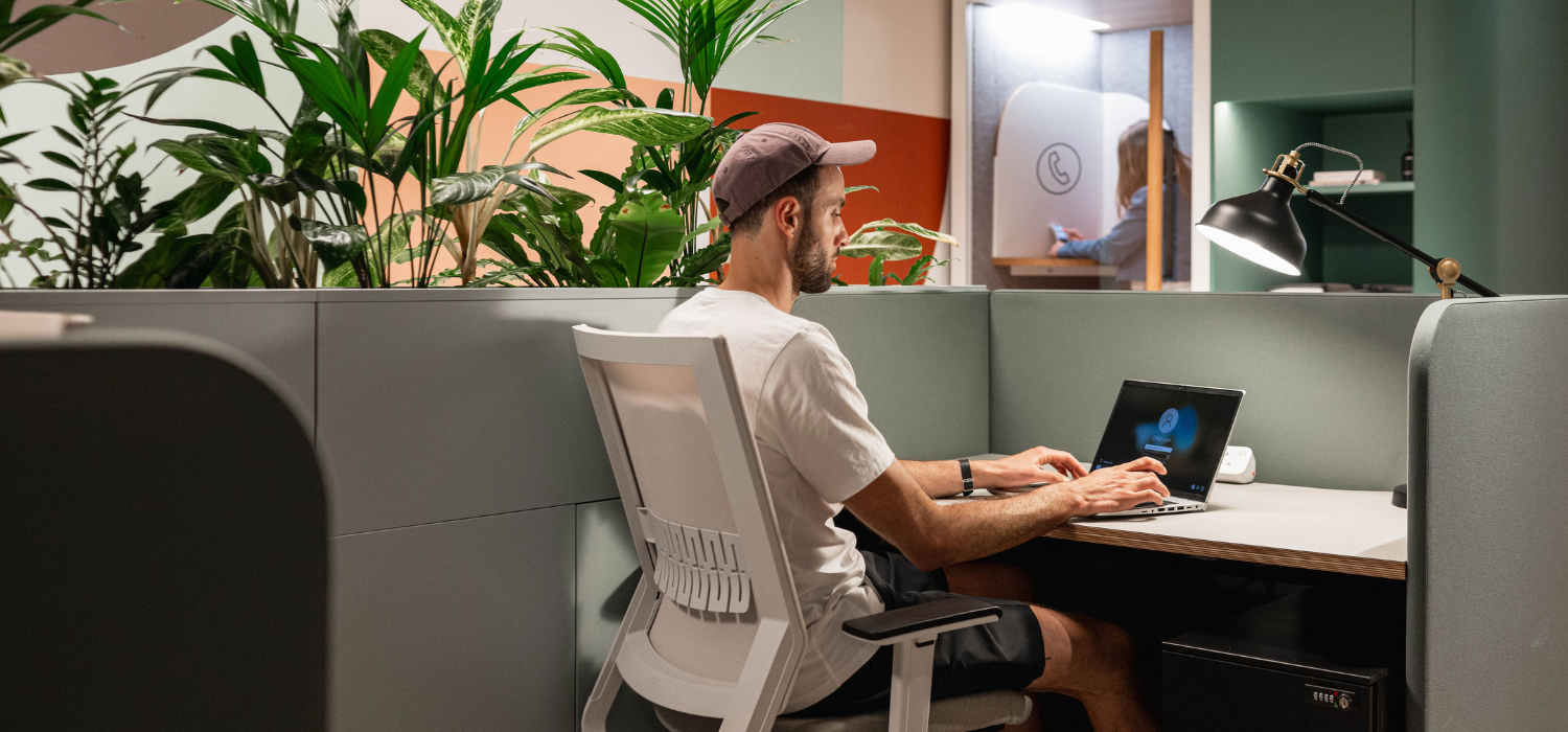 Une personne portant une chemise blanche et une casquette travaille sur un ordinateur portable à un bureau dans un espace de travail. La zone est entourée de cloisons végétales. Une autre personne est vue entrant dans une pièce avec un symbole circulaire sur la porte. L'éclairage est clair et naturel.