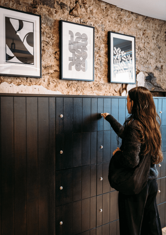 Une personne aux cheveux longs et noirs touche une armoire en bois sombre avec de petits boutons circulaires. Trois œuvres d'art abstraites encadrées sont accrochées sur un mur en pierre texturé au-dessus de l'armoire. La pièce dégage une atmosphère moderne et artistique.