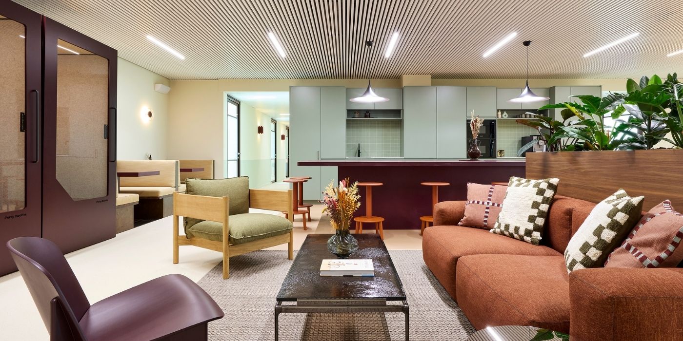 Salon moderne avec un canapé marron, un fauteuil vert, une table basse et des plantes. À l'arrière-plan se trouve une cuisine avec des armoires bleu clair et des tabourets de bar, ainsi que des cabines et des lampes suspendues.