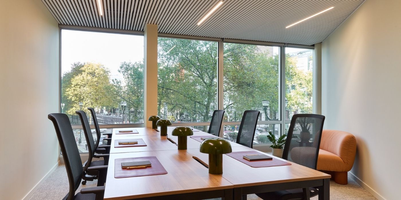 Salle de réunion de bureau moderne avec une longue table en bois, six chaises noires en maille, des lampes de bureau vertes, des blocs-notes et de grandes fenêtres laissant apparaître des arbres à l'extérieur. Une chaise orange rembourrée se trouve dans un coin.