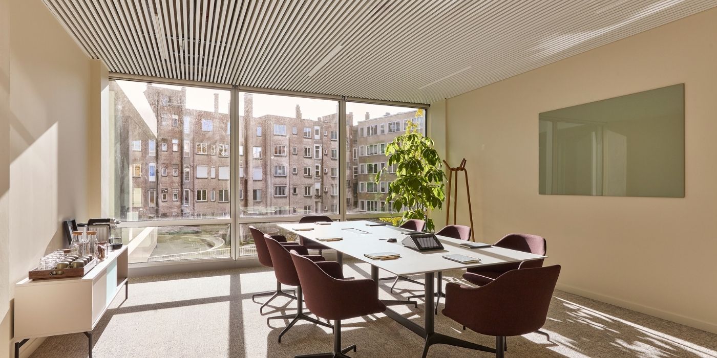 Une salle de réunion moderne avec une grande table, six chaises, une plante verte, une paroi vitrée avec vue sur la ville, un tableau blanc et un buffet avec des boissons. La lumière du soleil pénètre par les grandes fenêtres.