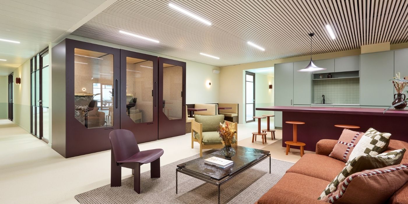 Salon de bureau moderne avec un canapé marron, une chaise violette, une table basse, des sièges de bar et des salles de réunion vitrées ; les tons neutres et le décor minimaliste créent une atmosphère confortable et professionnelle.