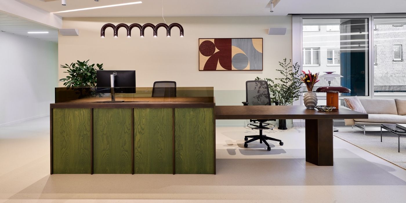 Espace de réception moderne avec un bureau en bois vert, deux chaises de bureau, un écran d'ordinateur, des plantes d'intérieur, des œuvres d'art murales abstraites et de grandes fenêtres laissant entrer la lumière naturelle.