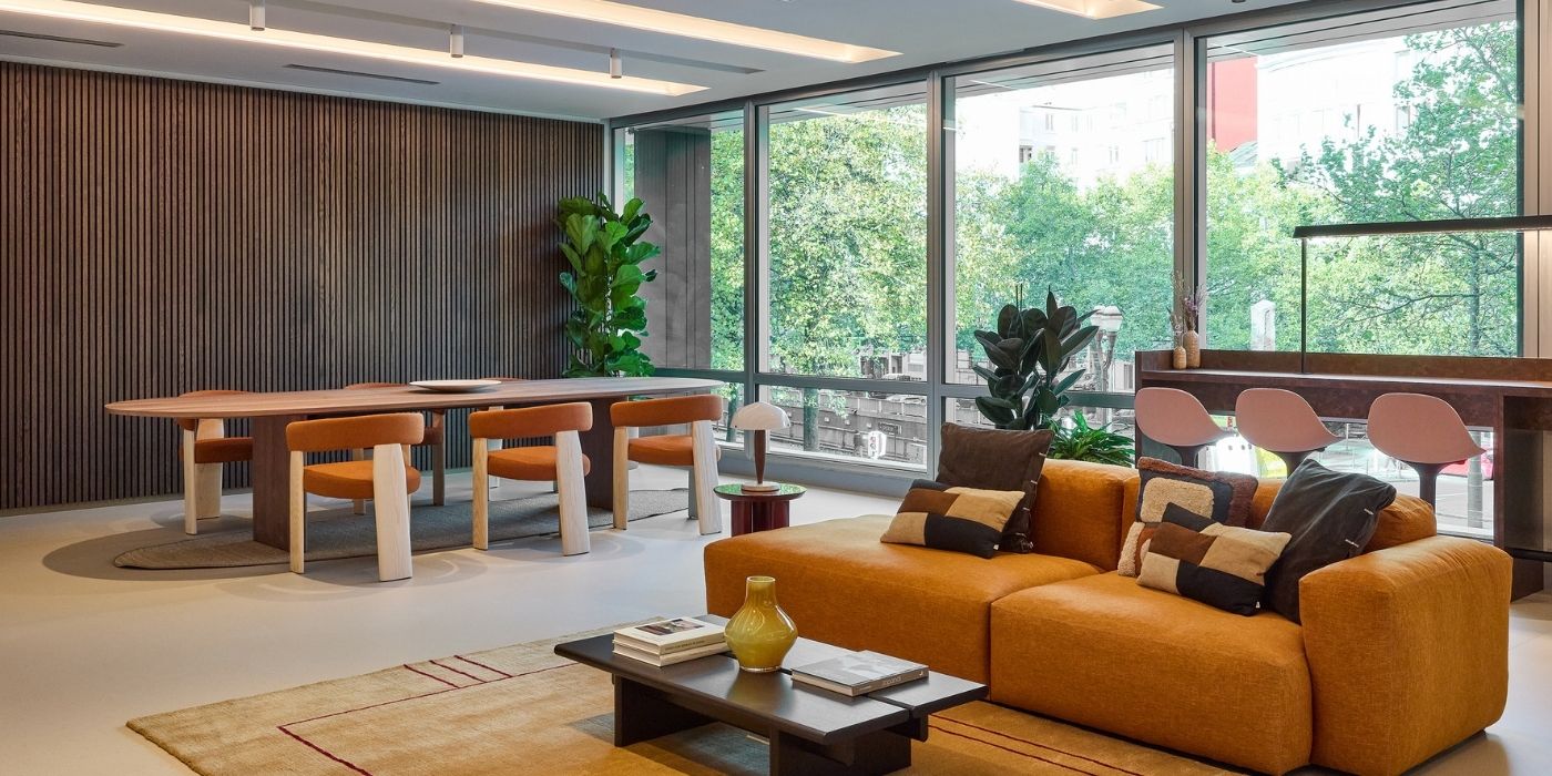 Un salon et une salle à manger modernes avec de grandes fenêtres, un canapé jaune, une table à manger avec six chaises orange, des plantes d'intérieur et des tabourets de bar sur un comptoir. La lumière naturelle remplit la pièce, qui donne sur les arbres verts à l'extérieur.