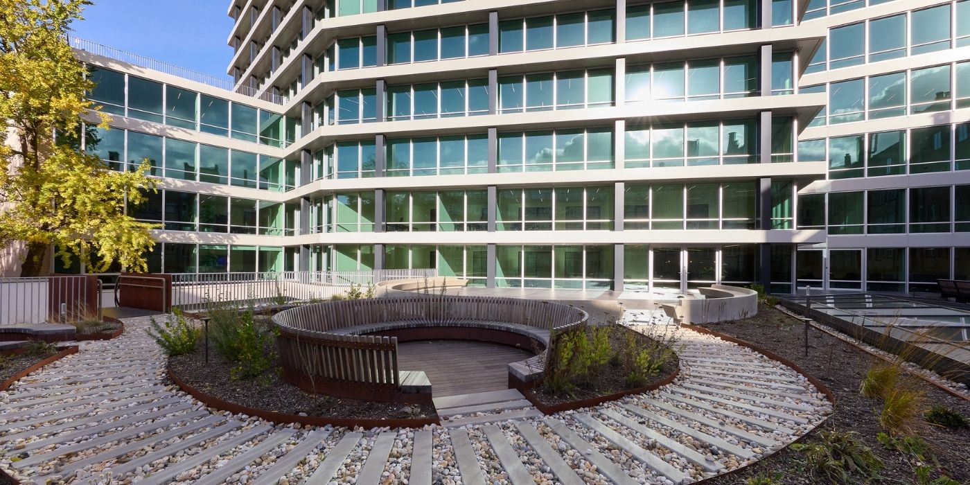 Ein modernes gläsernes Bürogebäude mit mehreren Stockwerken blickt auf einen begrünten Innenhof mit runden Holzbänken, Kieswegen und kleinen Pflanzen. Das Sonnenlicht wird von den Fenstern reflektiert.