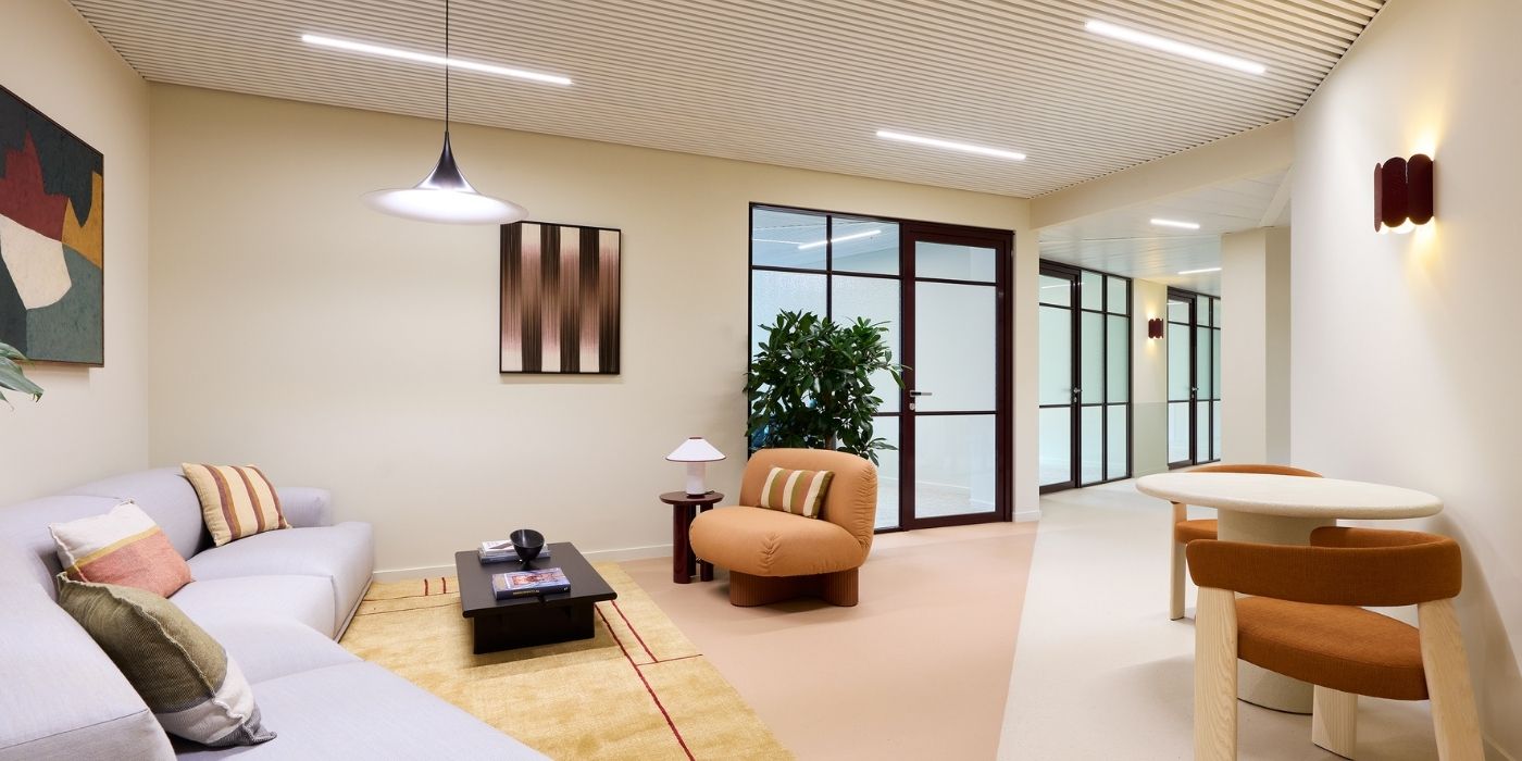 Salon moderne composé d'un canapé gris clair, d'un fauteuil rond de couleur bronze, d'une petite table basse et d'un ensemble de salle à manger avec deux chaises orange. La grande fenêtre, la plante d'intérieur, l'art abstrait et l'éclairage doux créent une atmosphère confortable et élégante.