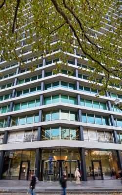 Un grand immeuble de bureaux moderne avec des fenêtres en verre et une façade géométrique, vu de la rue. Des personnes marchent sur le trottoir en contrebas, et des branches d'arbres aux feuilles vertes encadrent le haut de l'image.