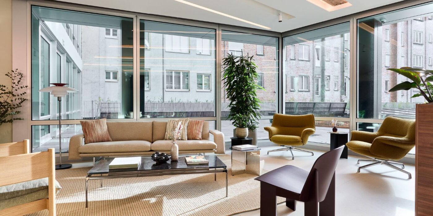 Un salon moderne avec de grandes fenêtres allant du sol au plafond, un canapé beige, deux fauteuils moutarde, une table basse noire, des plantes et les bâtiments de la ville visibles à l'extérieur. L'espace est baigné de lumière naturelle.