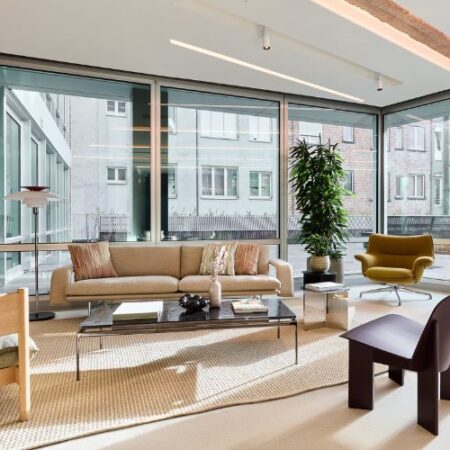 A modern living room with large floor-to-ceiling windows, a beige sofa, green armchair, wooden chairs, a black coffee table, neutral rug, and a tall potted plant. Urban buildings are visible outside.