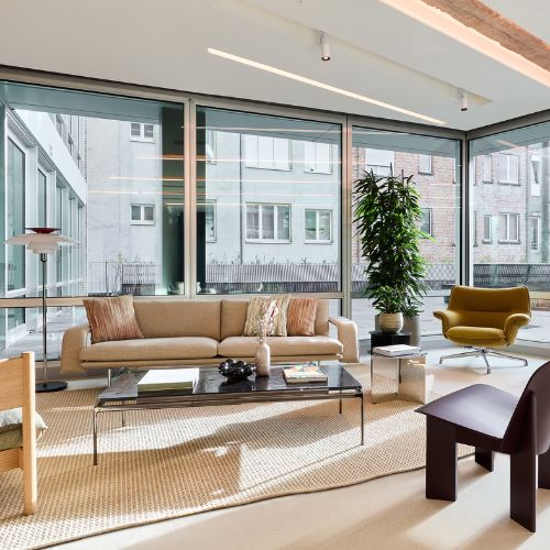 Een moderne woonkamer met grote ramen van vloer tot plafond, een beige bank, groene fauteuil, zwarte salontafel, kamerplant en neutrale inrichting, waardoor natuurlijk licht de ruimte vult.