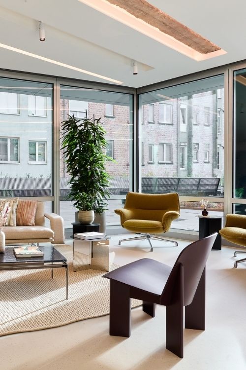 Un salon moderne avec de grandes fenêtres allant du sol au plafond, un canapé beige, deux fauteuils moutarde, une table basse noire, des plantes et les bâtiments de la ville visibles à l'extérieur. L'espace est baigné de lumière naturelle.