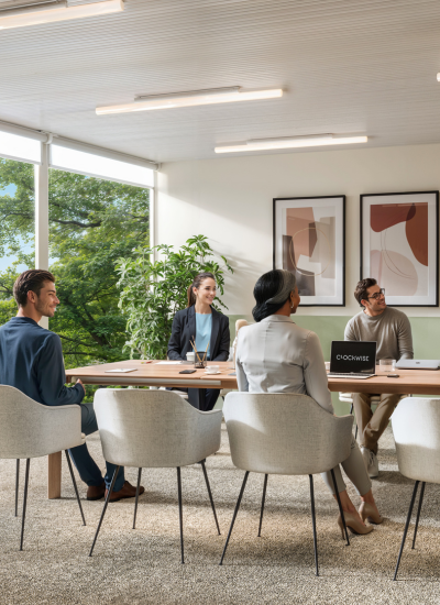 Quatre personnes sont assises autour d'une table de conférence dans un bureau moderne et lumineux, avec de grandes fenêtres et des œuvres d'art abstraites aux murs, en réunion. Un ordinateur portable et des documents sont posés sur la table.