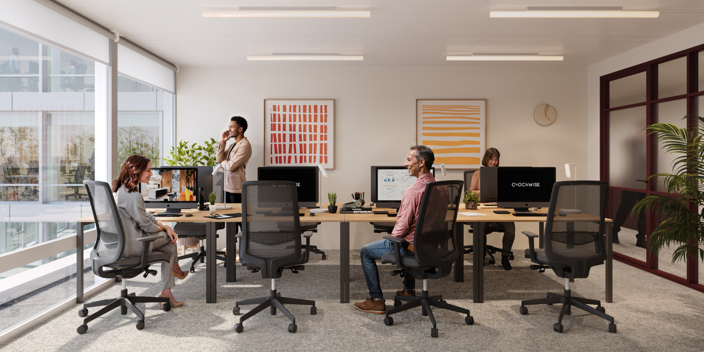 Bureau ouvert moderne avec quatre personnes travaillant à des bureaux, des ordinateurs et des plantes sur des tables, une grande fenêtre et des œuvres d'art abstrait colorées au mur. Deux personnes sont assises, une personne debout au téléphone et une autre travaille sur un ordinateur.