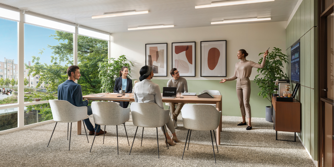 Une femme se lève et présente à quatre personnes assises à une table de conférence dans un bureau moderne avec de grandes fenêtres, des plantes et de l'art abstrait sur les murs.
