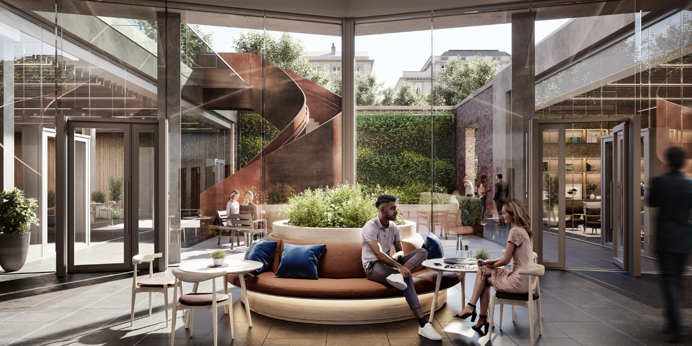 Deux personnes sont assises et discutent sur un banc circulaire avec une jardinière dans un atrium moderne et ensoleillé. De grandes fenêtres, de la verdure et un escalier en colimaçon spectaculaire créent une atmosphère ouverte et accueillante. D'autres personnes sont présentes à l'arrière-plan.