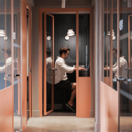 A man sits in a small, glass-walled office booth, working on a laptop. The booth is painted in warm tones and illuminated by a wall-mounted lamp. The hallway outside the booth is visible through the glass.