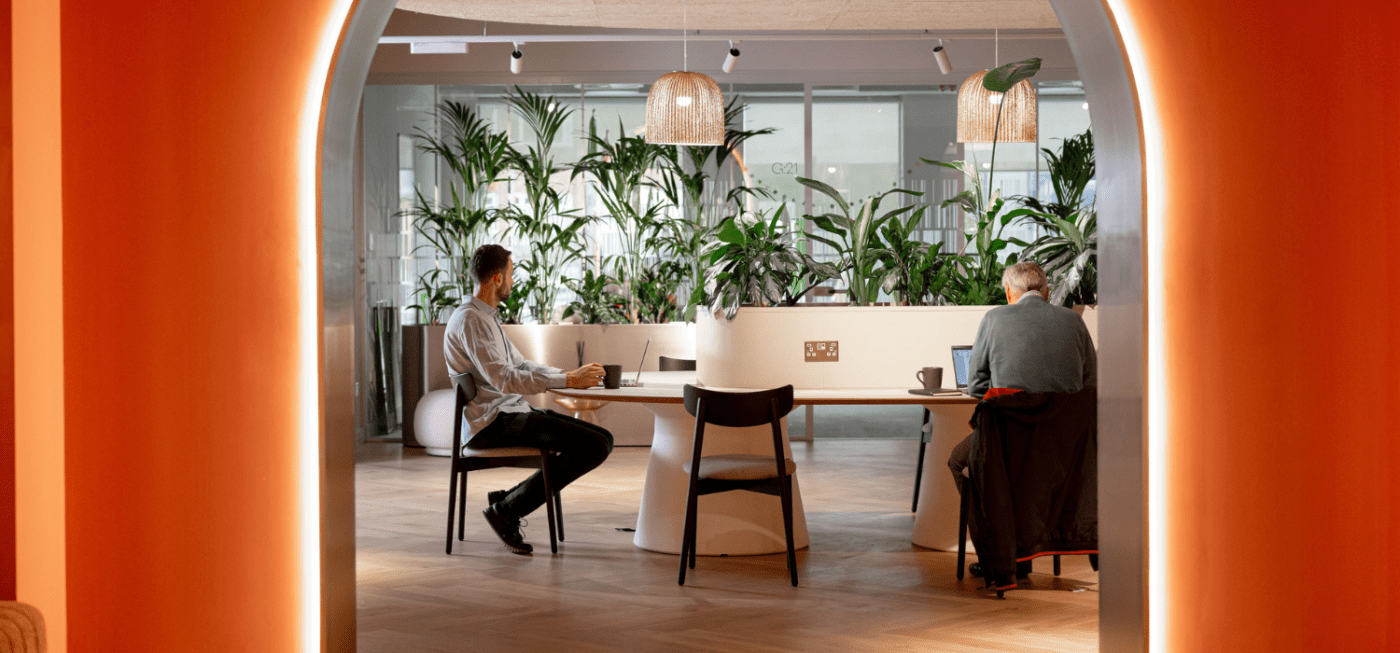 A modern office space with two people sitting at a long table, engaged in work on laptops. The setting includes lush green plants in the background, wooden flooring, and hanging woven lamps, viewed through an arched doorway.