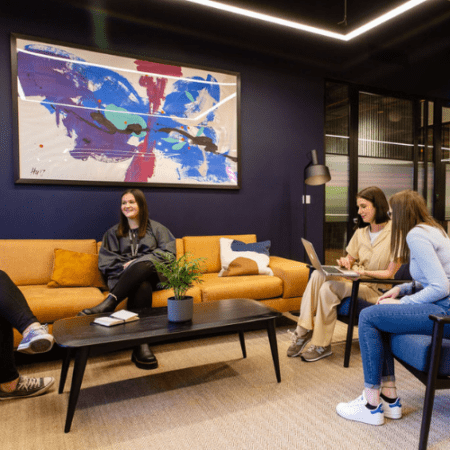 Four people are sitting and talking in a modern office lounge. The space has a yellow sofa, a black coffee table, a potted plant, and a large abstract painting on a dark wall. Two people use a laptop, and one person has a notebook.