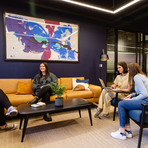 Four people are sitting and talking in a modern office lounge. The space has a yellow sofa, a black coffee table, a potted plant, and a large abstract painting on a dark wall. Two people use a laptop, and one person has a notebook.