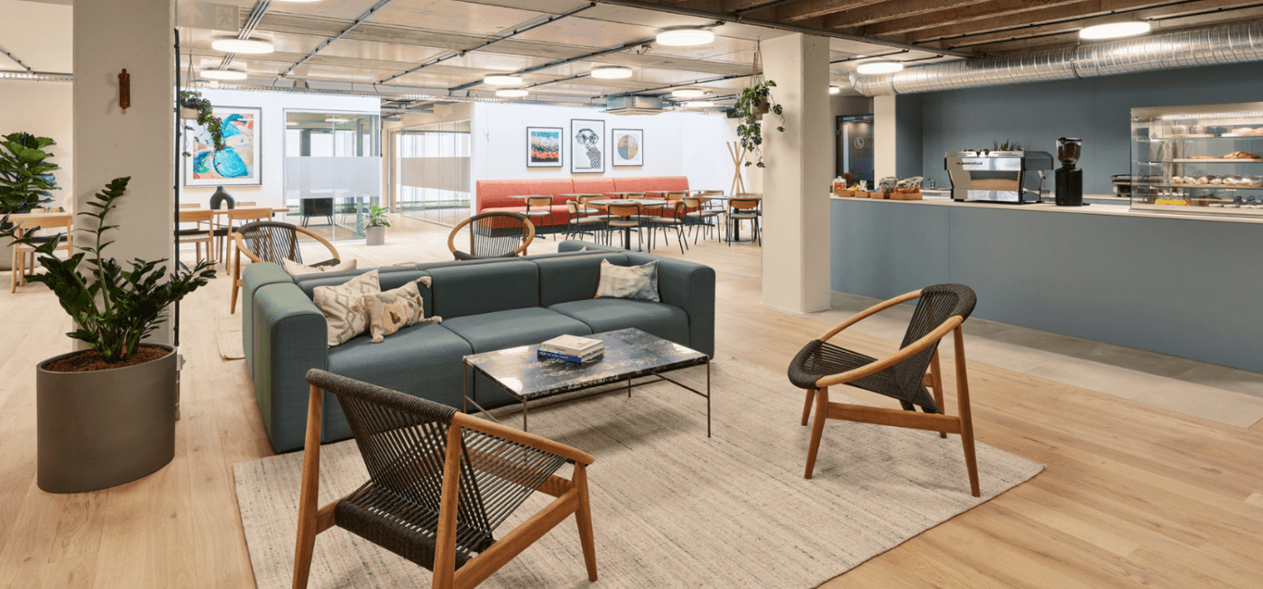 Modern office lounge with a teal sofa, two chairs, and a coffee table on a rug. Open layout includes bar area with snacks and coffee machine. Dining tables and potted plants enhance the contemporary decor. Artworks adorn the walls in the background.