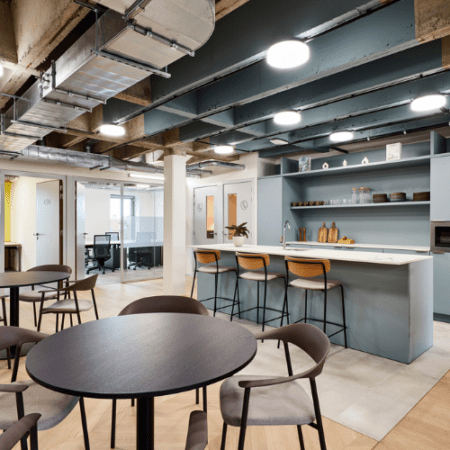 Modern office kitchen area featuring a long counter with four stools, open shelving with various dishes, and a seating area with round tables and chairs. The space has exposed ductwork and soft lighting, creating an industrial yet inviting atmosphere.