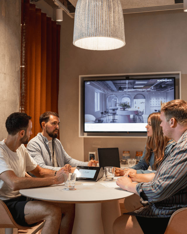 Four people sit around a table engaged in discussion. A digital tablet and laptop are on the table. A large screen behind them displays an interior design image. The setting appears to be an office or meeting room with a modern aesthetic.