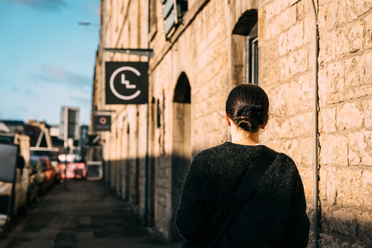 Person with dark hair in a bun, wearing a dark sweater, walks down a sunlit stone street. Black sign with a logo hangs from a building. Cars line the left side of the street under a clear blue sky.