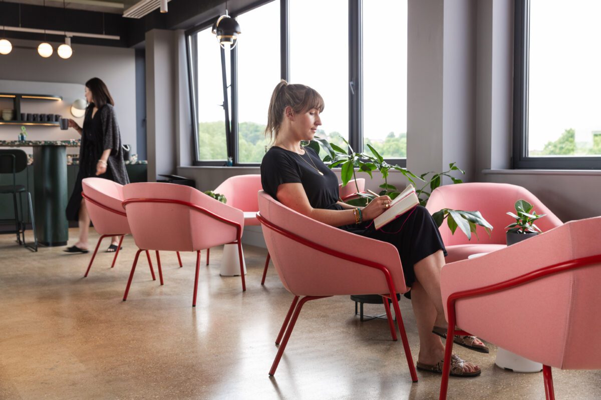 A woman sits in a modern office lounge on a pink chair, writing in a notebook. The room has large windows, several pink chairs, and potted plants. Another person is walking in the background. The atmosphere is bright and airy.